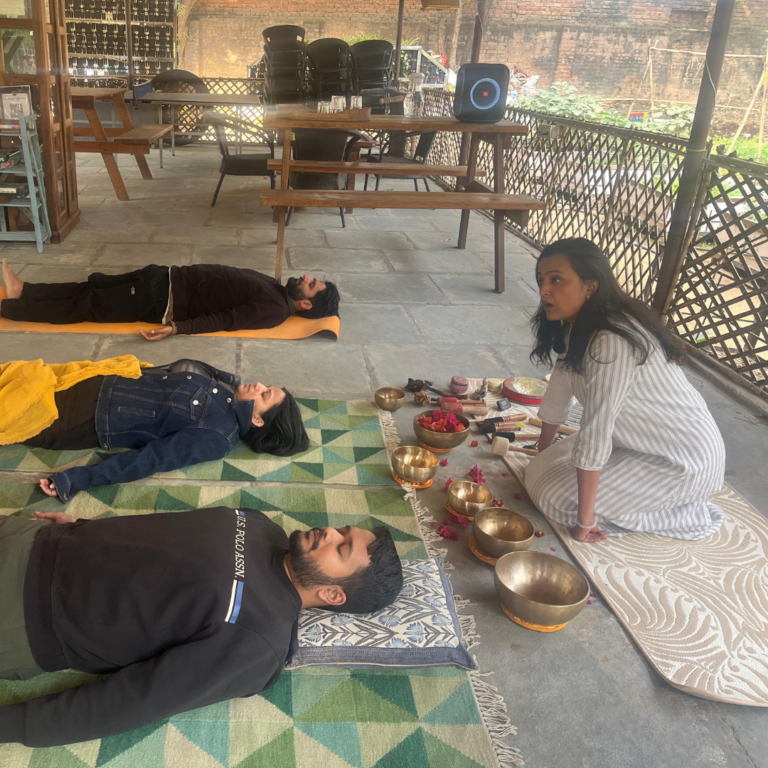 A sound therapist guiding a group during a sound therapy session with participants lying on mats surrounded by Tibetan singing bowls.