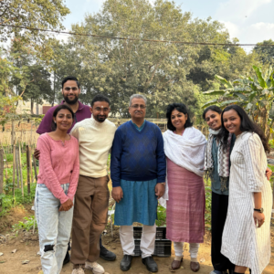 A group of seven people posing together outdoors in a lush green garden under a bright sky.