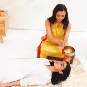 A woman performing sound healing therapy with a Tibetan singing bowl on a person lying peacefully in a bright and serene room.