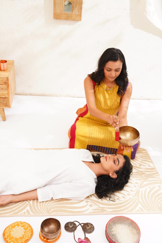 A woman performing sound healing therapy with a Tibetan singing bowl on a person lying peacefully in a bright and serene room.